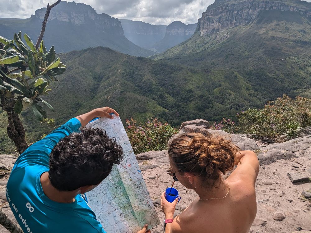 A local guide in a blue shirt and a hiker examining a trail map together while resting at a scenic valley viewpoint. 