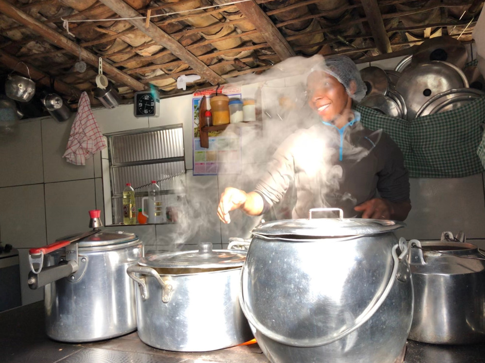 A local woman smiling as she cooks a traditional homemade meal using large aluminum pots over a stove. 