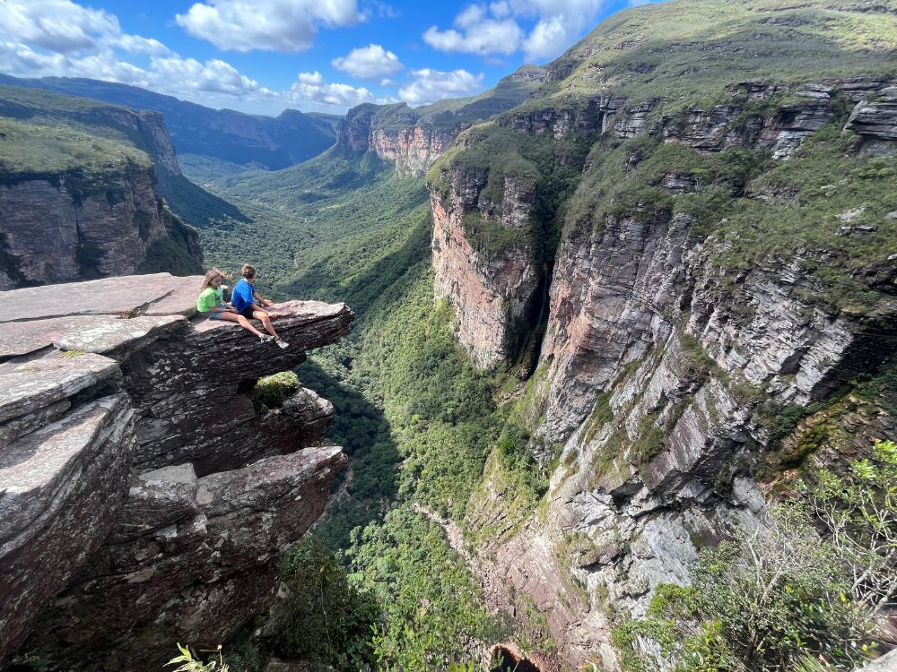 Two people sitting on the edge of a dramatic 300-meter-high canyon cliff at Cachoeirão, admiring the steep rocky walls. 