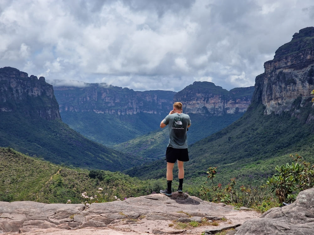  A hiker standing on a rocky outcrop taking a photograph of the sprawling green valleys and flat-topped mountains.
