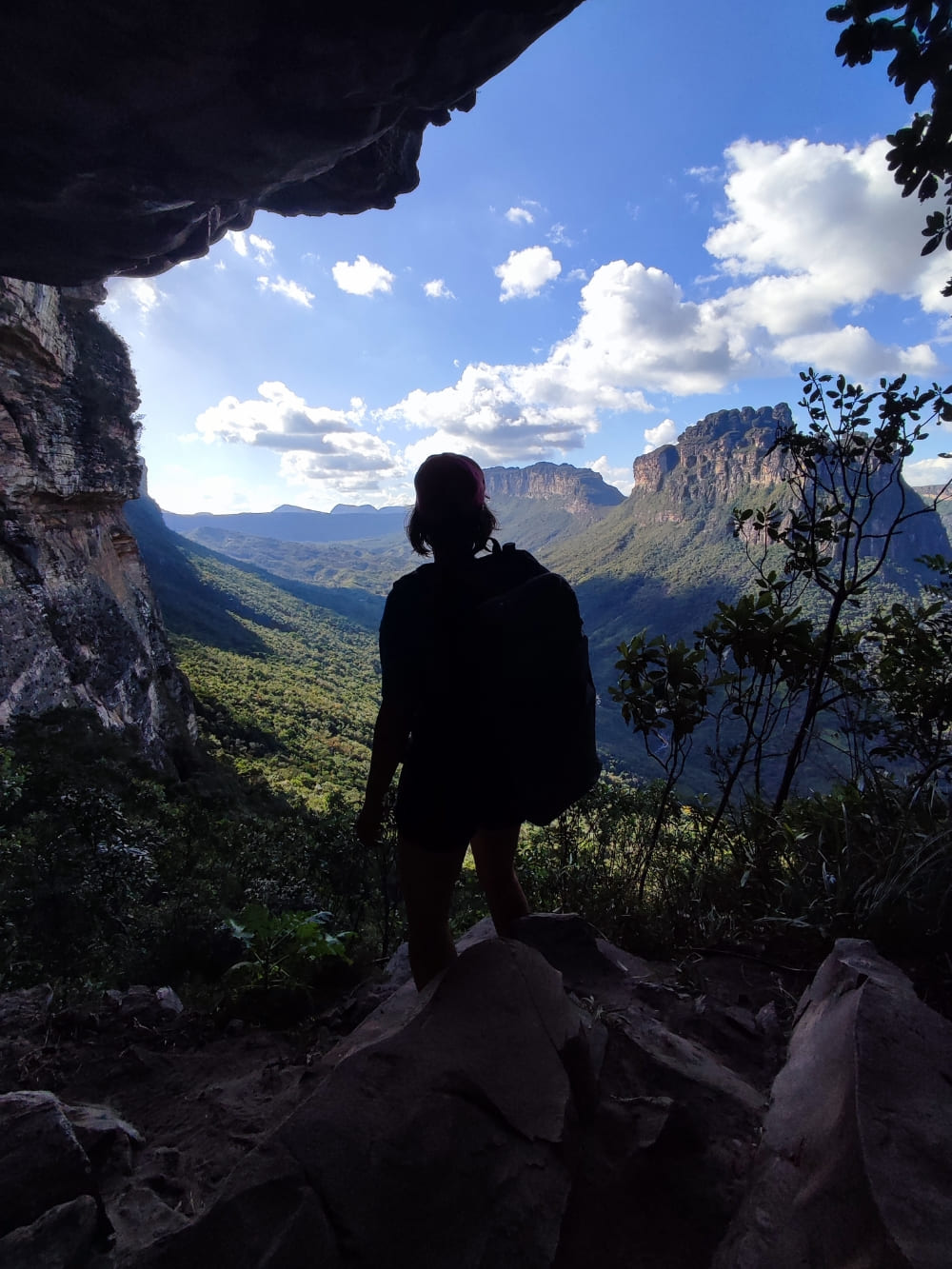 Silhouette of a hiker wearing a backpack standing at a dark cave entrance, looking out over the sunlit Pati Valley. 