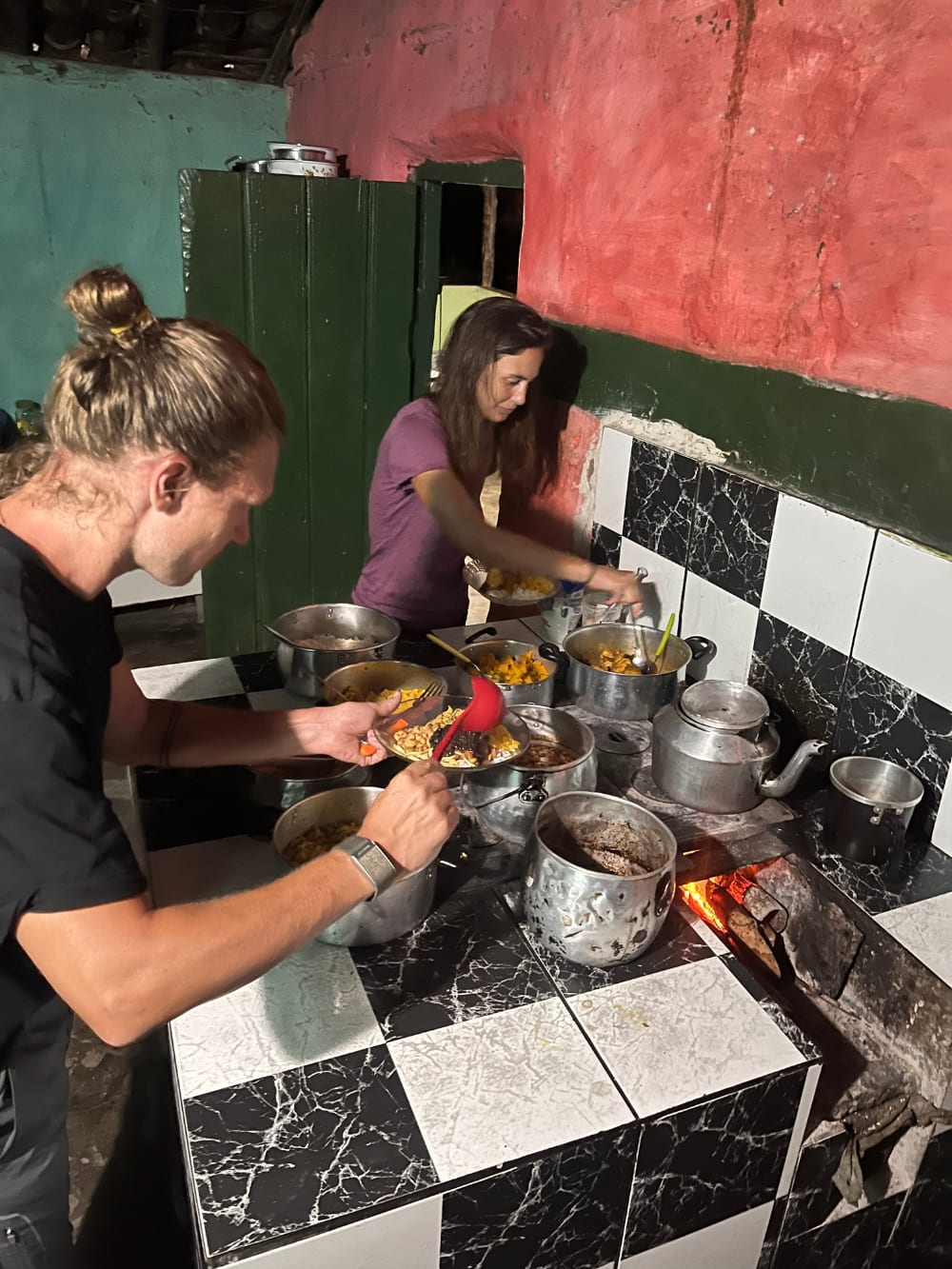 Two hikers serving themselves fresh homemade food from various pots in a rustic homestay kitchen.