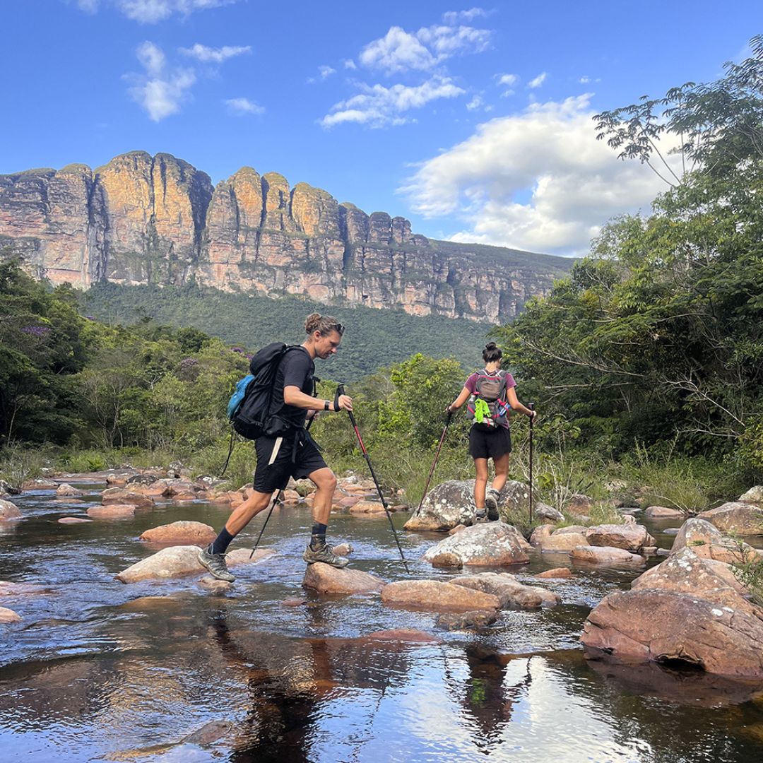 Two hikers using trekking poles to cross a shallow, rocky river with a large mountain peak towering in the background. 