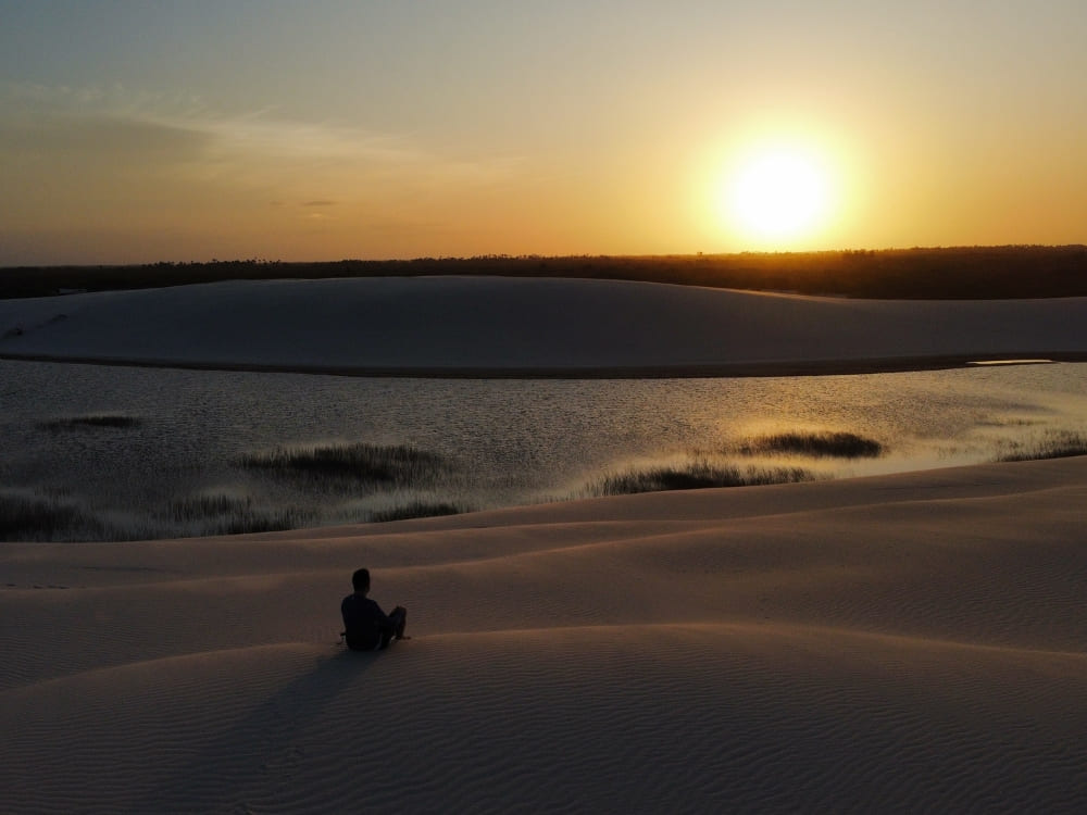 Silhouette of a person sitting on a sand dune, watching a golden sunset over the lagoons of Lençóis Maranhenses. Silhouette of a person sitting on a sand dune, watching a golden sunset over the lagoons of Lençóis Maranhenses.
