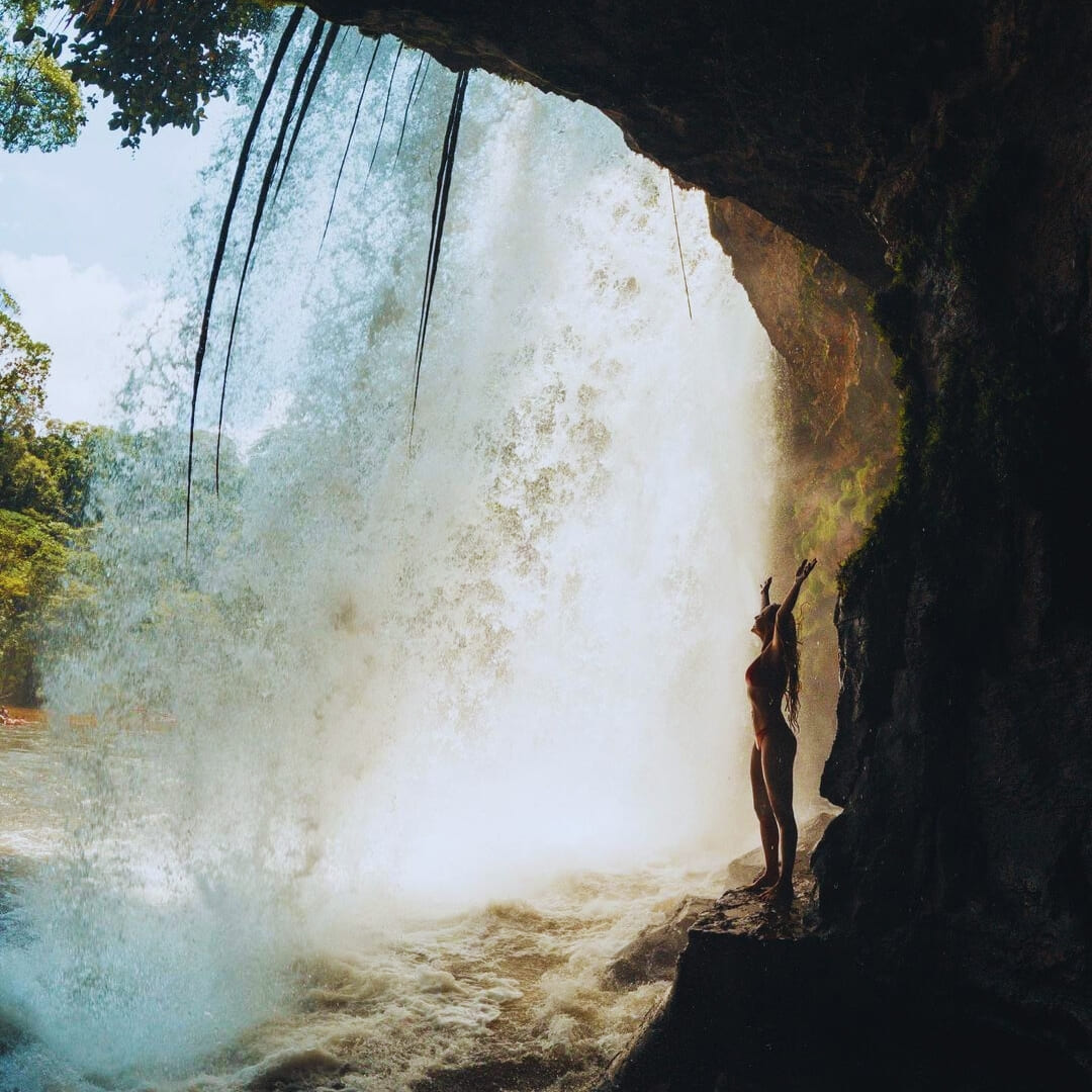A traveler stands behind a powerful rushing waterfall in the Amazon Rainforest with their arms raised. A traveler stands behind a powerful rushing waterfall in the Amazon Rainforest with their arms raised.