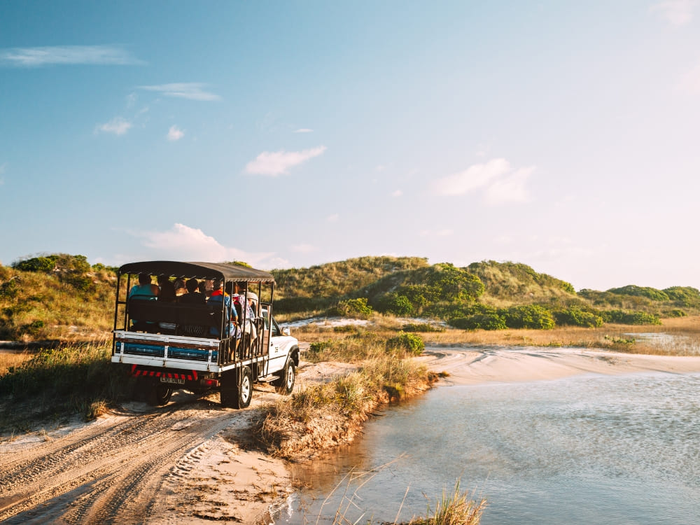 Tourists in an adapted 4x4 vehicle drive along a sandy water trail, illustrating the transport options for exploring the Lençóis Maranhenses lagoons.