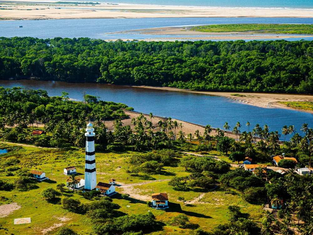 Aerial view of a tall black and white striped lighthouse and lush green vegetation near the base cities for Lençóis Maranhenses tours. Aerial view of a tall black and white striped lighthouse and lush green vegetation near the base cities for Lençóis Maranhenses tours.