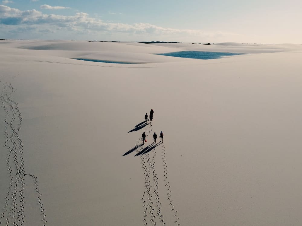 Aerial view of a group trekking across the immense white sand dunes of Lençóis Maranhenses, with small blue lagoons in the background.