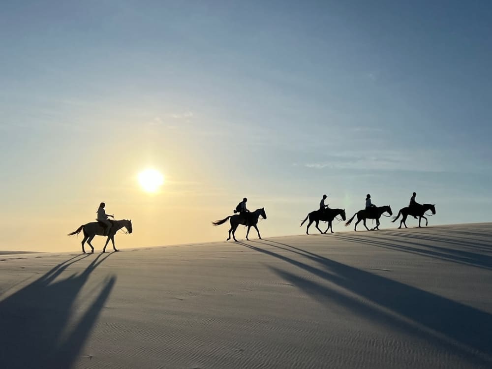 Silhouettes of five people horseback riding across the sand dunes of Lençóis Maranhenses under the bright late afternoon sun.