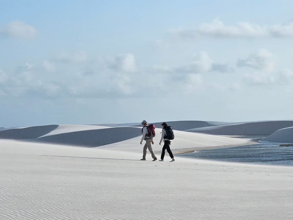 Two backpackers hike across the vast white sand dunes next to a freshwater lagoon in Lençóis Maranhenses National Park.