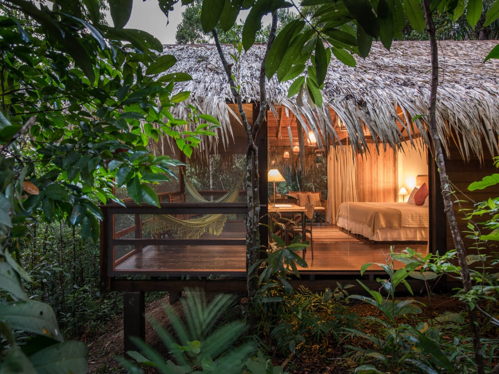 A luxury thatched-roof bungalow with warm interior lighting sits nestled among lush green trees at the Anavilhanas Jungle Lodge.