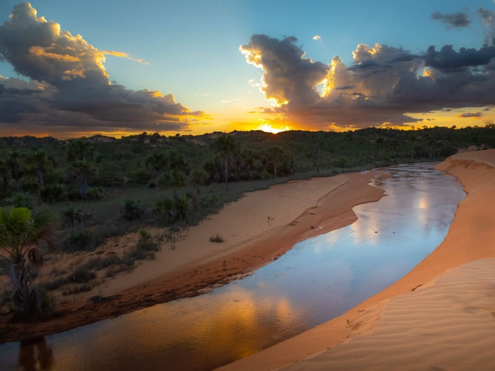 A calm river flowing through wide sandy areas and rugged cliffs during sunset, reflecting the region's geological past 