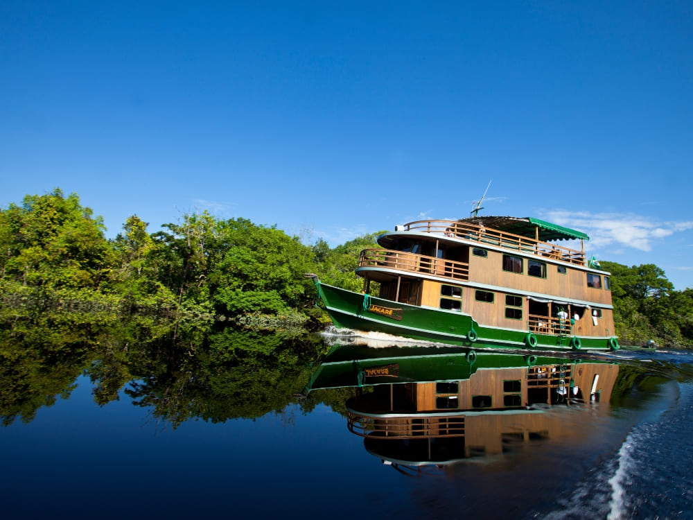 A rustic, three-deck wooden riverboat cruises smoothly along a dark Amazonian river beneath a blue sky. 