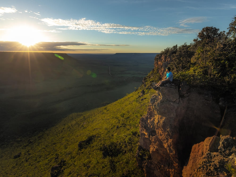 A traveler sits on a cliff edge enjoying the sunset over the vast savanna landscape of Jalapão State Park.