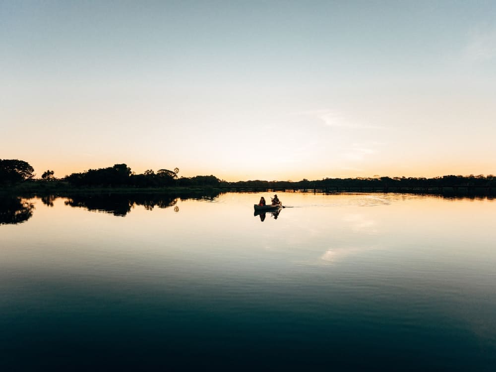 Two people paddling a canoe on a calm river that mirrors the sky in the area where is the Pantanal. Two people paddling a canoe on a calm river that mirrors the sky in the area where is the Pantanal.