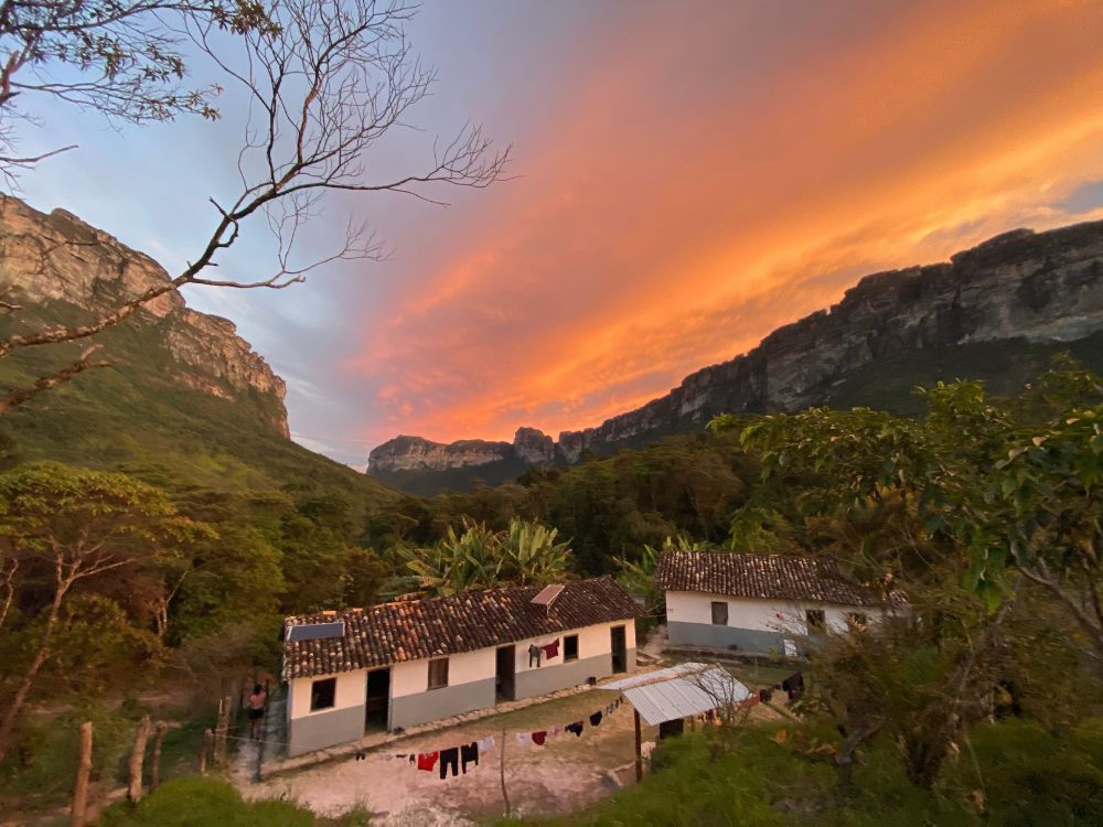 Modest white homestay houses with clay roofs nestled in a green valley beneath towering mountains, under a dramatic orange sunset sky. Modest white homestay houses with clay roofs nestled in a green valley beneath towering mountains, under a dramatic orange sunset sky.