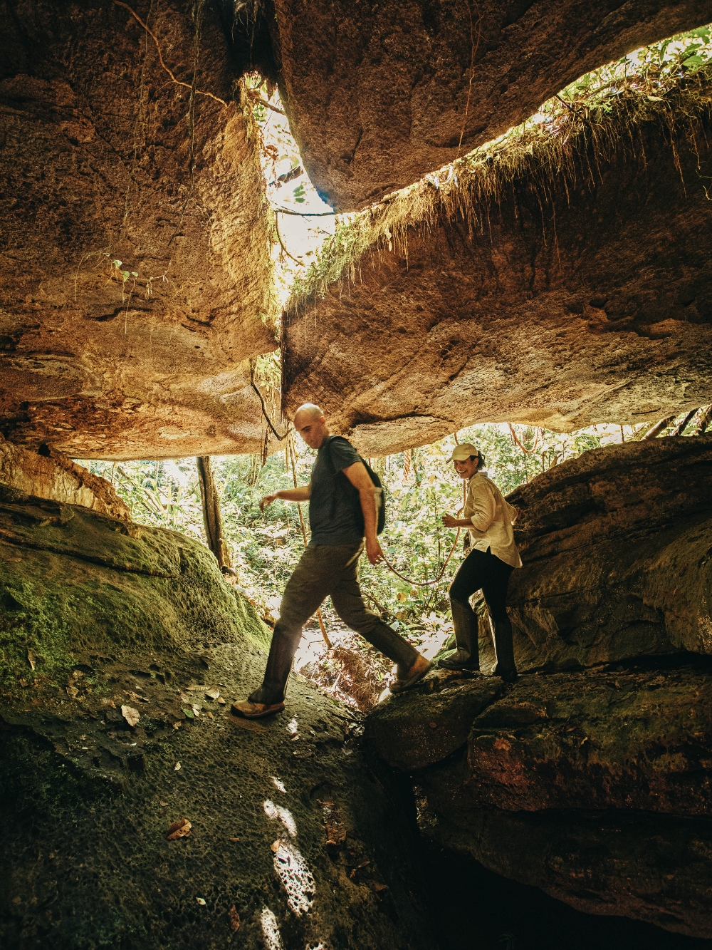 Two travelers hiking under a massive rock formation in the Presidente Figueiredo forest.