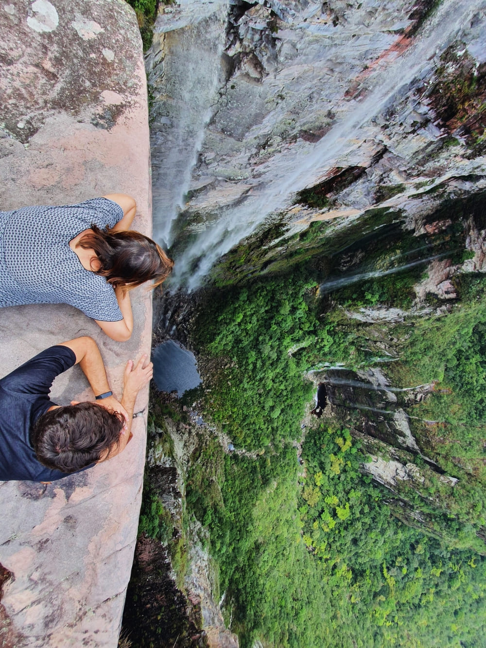 Two hikers look down a steep cliff edge at a lush green valley while trekking the iconic Vale do Pati route. 