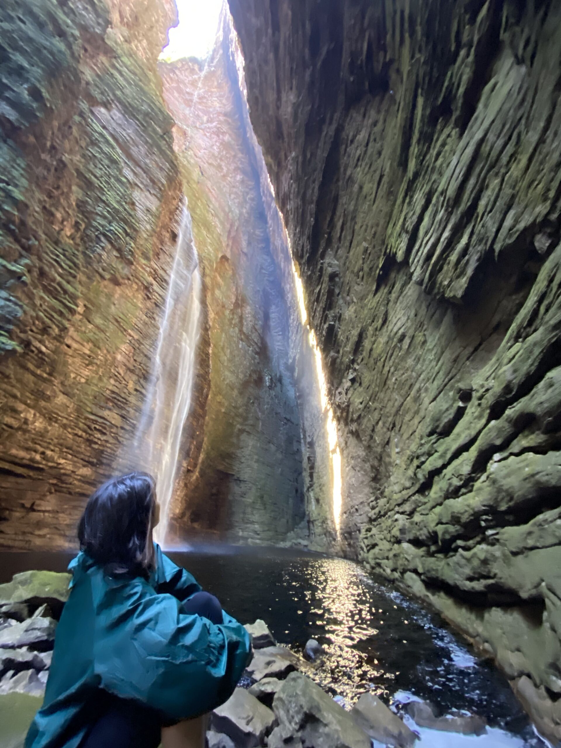 A hiker in a green jacket admires a towering waterfall inside a steep canyon during the rainy season in Chapada Diamantina. 