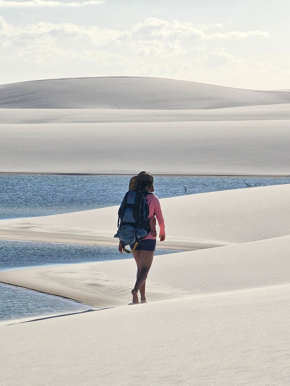 A traveler carrying a backpack walks barefoot along the shoreline of a peaceful lagoon in Santo Amaro.
