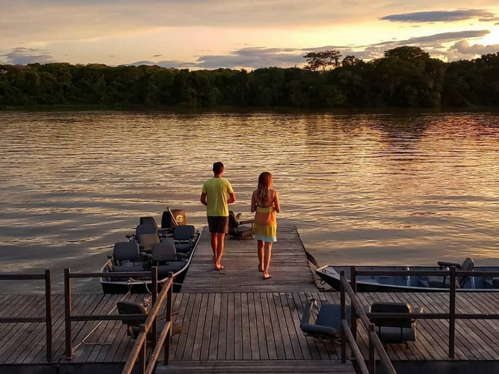 A couple walks down a wooden pier toward safari boats during a beautiful sunset over a Pantanal river. A couple walks down a wooden pier toward safari boats during a beautiful sunset over a Pantanal river.