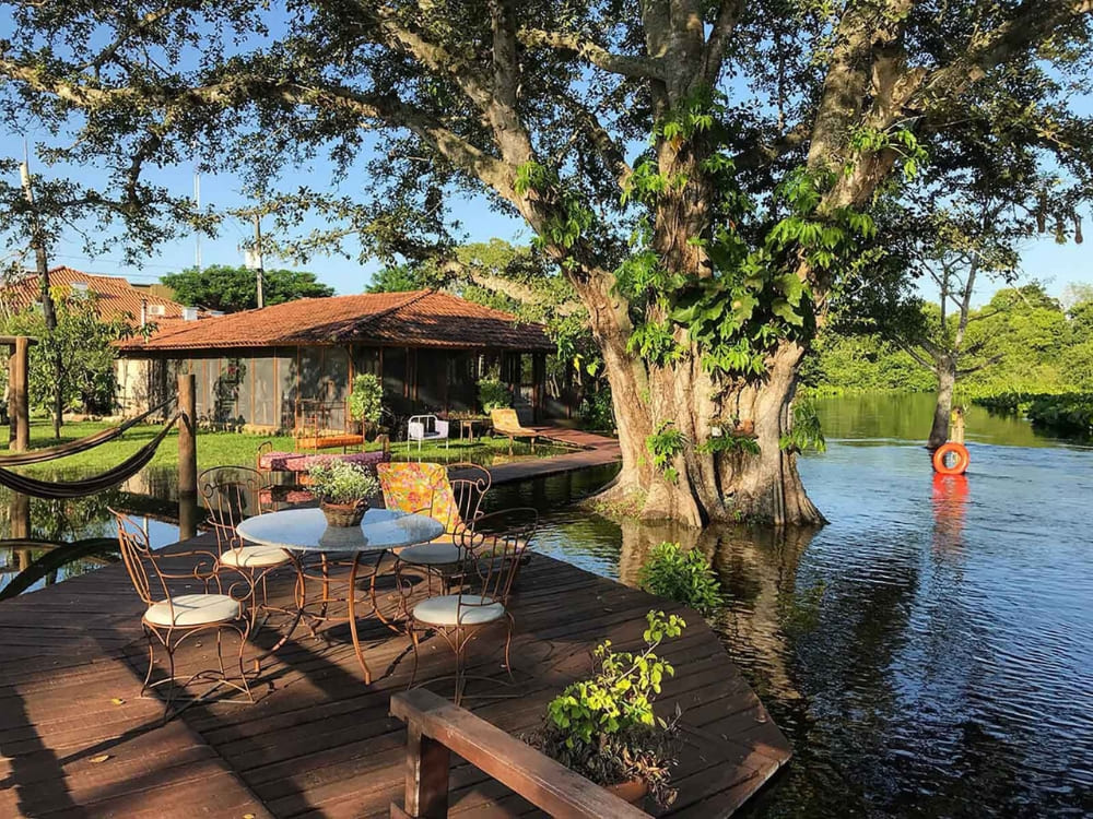 A wooden deck with a table and chairs sits beside a tranquil lake and a massive tree at RefĂşgio da Ilha ecolodge. A wooden deck with a table and chairs sits beside a tranquil lake and a massive tree at RefĂşgio da Ilha ecolodge.