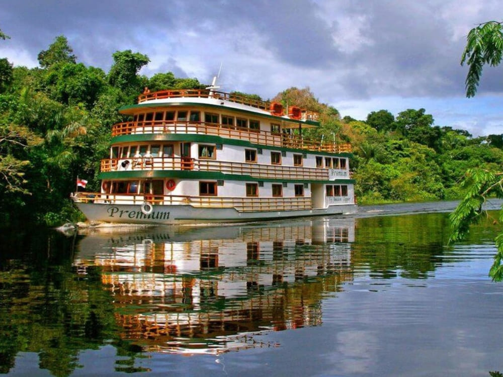 The Amazon Clipper Premium riverboat cruising along the Amazon River, reflecting in the calm water against a backdrop of dense green rainforest.