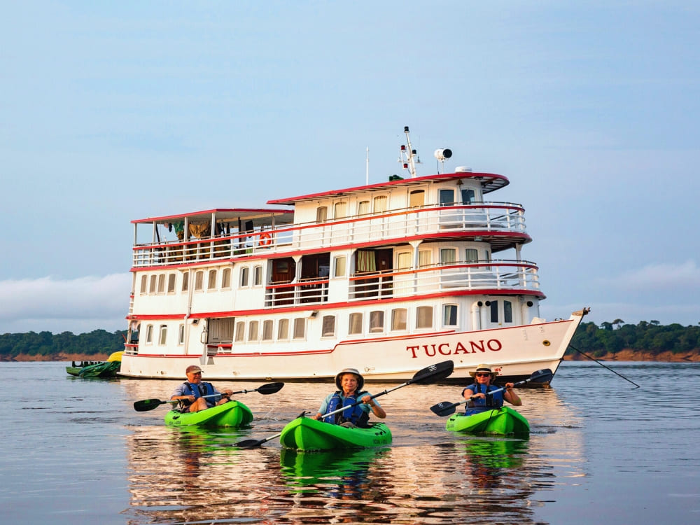 Three people paddle green kayaks on a calm river in front of a multi-deck riverboat named Tucano, with lush green trees lining the shore in the background.