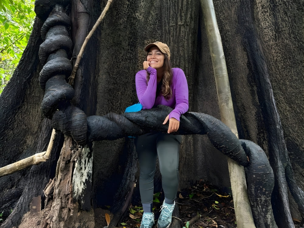 Smiling woman wearing a purple shirt, leaning against the thick, twisted roots of a giant tree in the middle of a lush green forest.