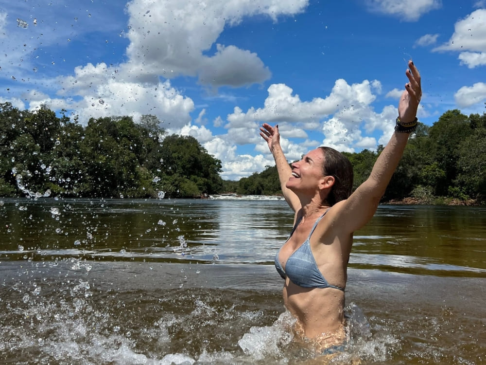 Smiling woman bathing in the calm waters of a river surrounded by green trees, splashing water into the air with her arms wide open.