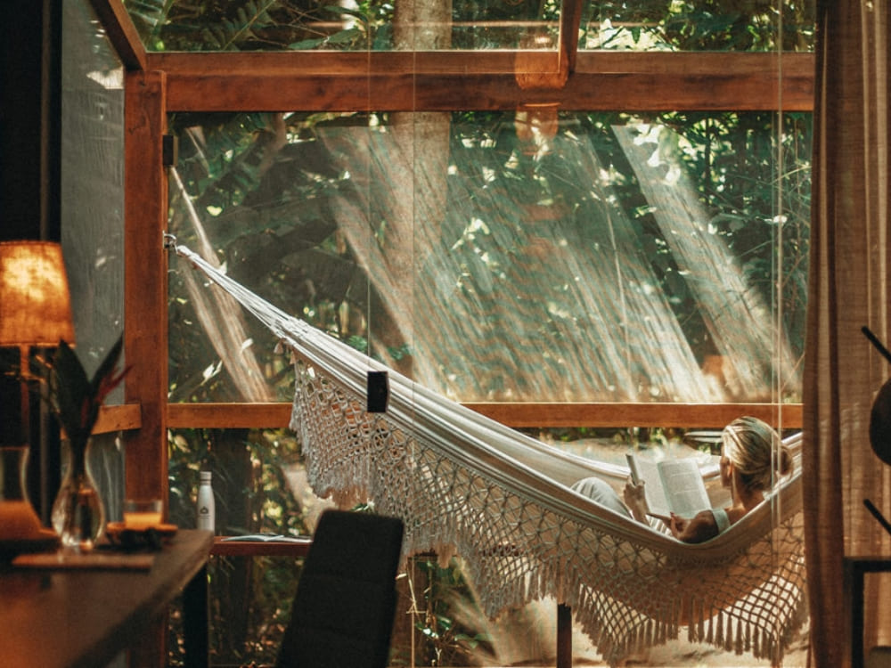 A person relaxing in a hammock inside a sophisticated room with large glass windows facing the jungle. A person relaxing in a hammock inside a sophisticated room with large glass windows facing the jungle.