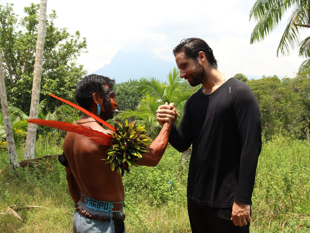 A Yanomami indigenous guide wearing traditional red feathers shaking hands with a hiker in the forest. 