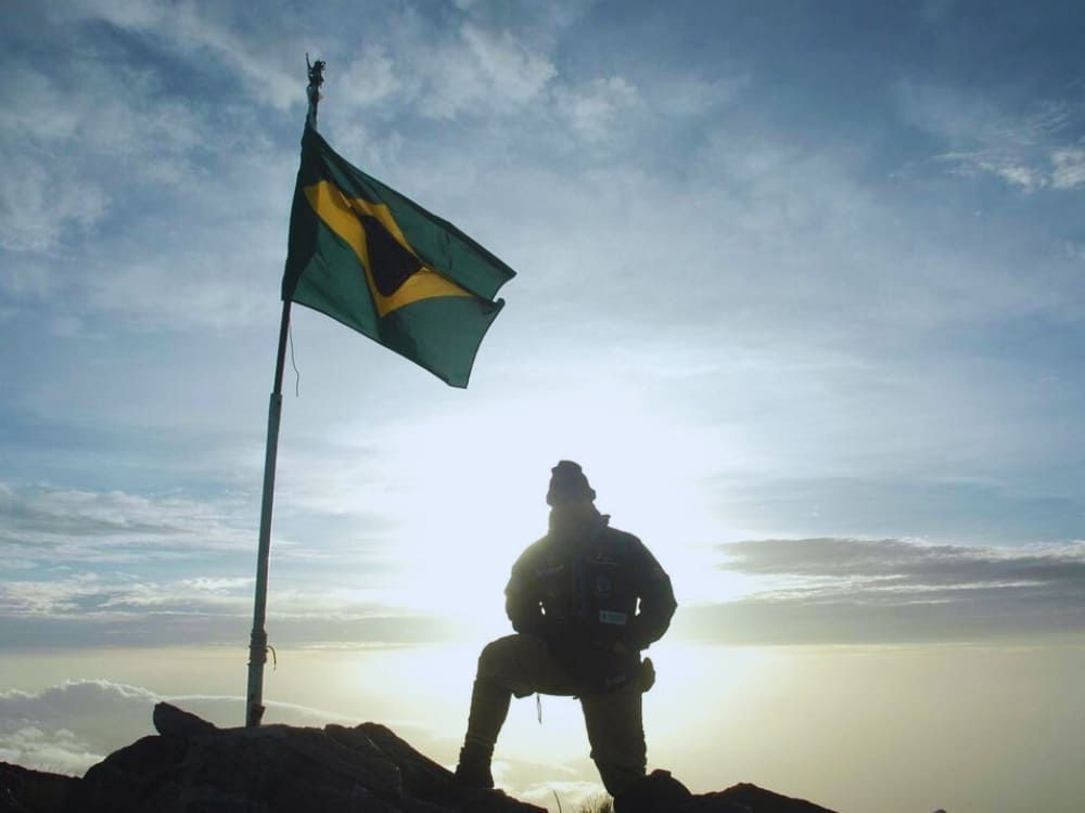 Silhouette of an explorer kneeling next to a waving Brazilian flag at the summit of Pico da Neblina at sunrise. 