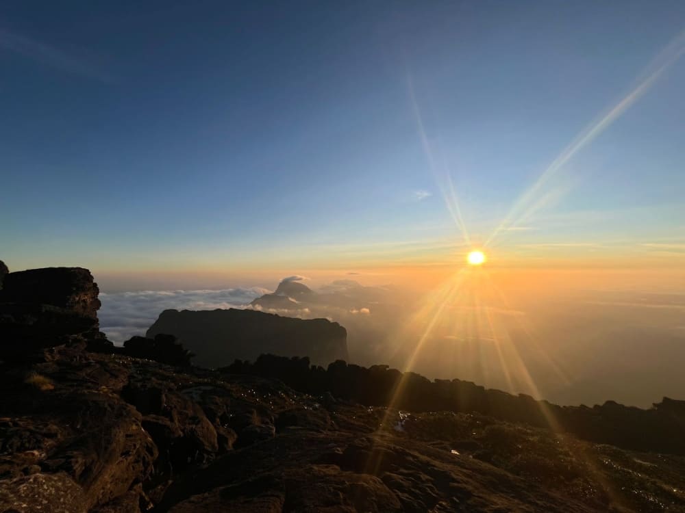 The sun bursts with bright rays over a sea of white clouds and silhouetted table mountains, viewed from the rocky summit of Mount Roraima.