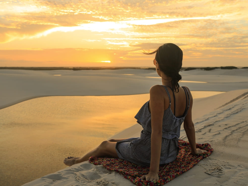 A woman sits on a patterned towel atop a dune, enjoying the warm Lençóis Maranhenses weather and watching the sunset over a serene lagoon. 