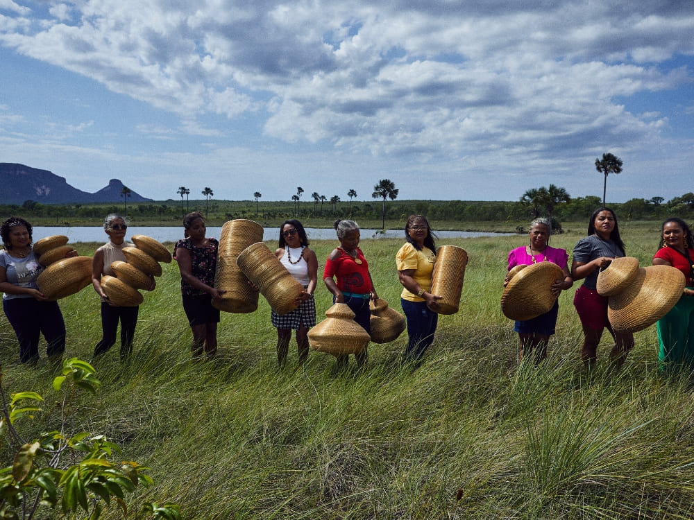  A group of women standing in a lush green field under a cloudy sky, holding up woven handicrafts made from Golden Grass in the Jalapão region.