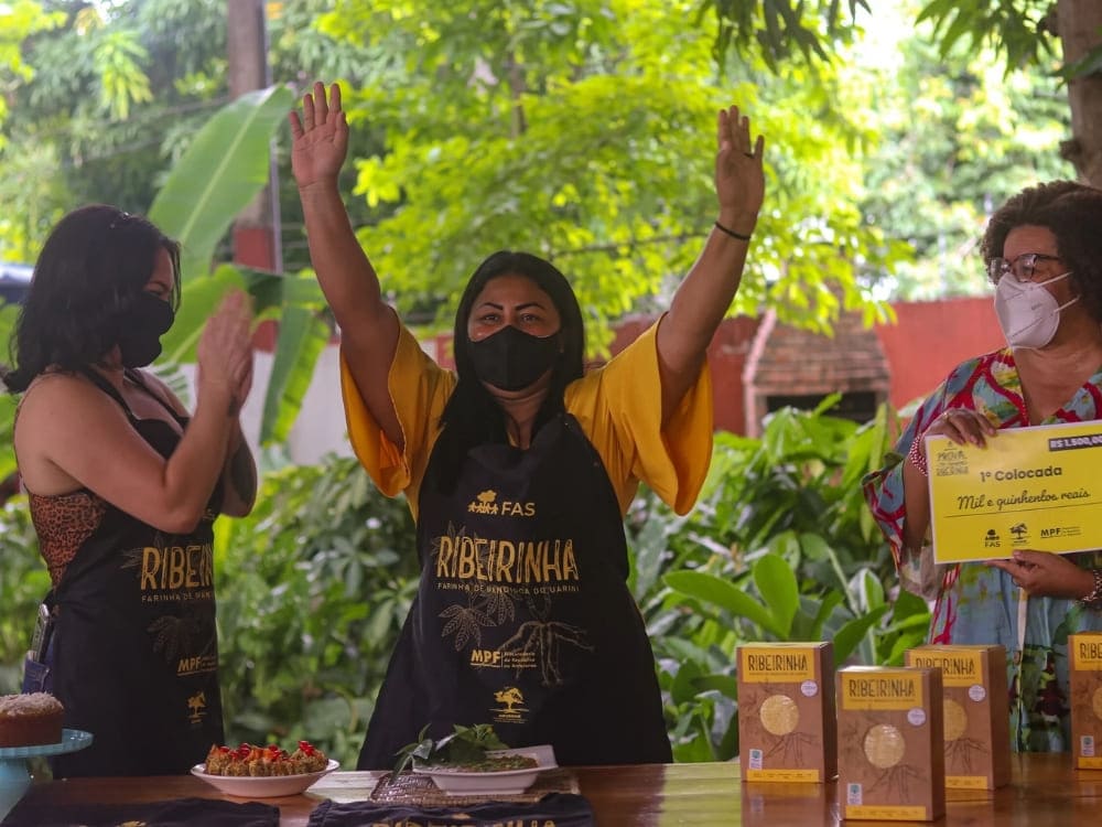 A woman wearing a black apron with FAS Ribeirinha branding raises her hands in joy, standing alongside others celebrating the Sustainable Amazon Foundation's program.