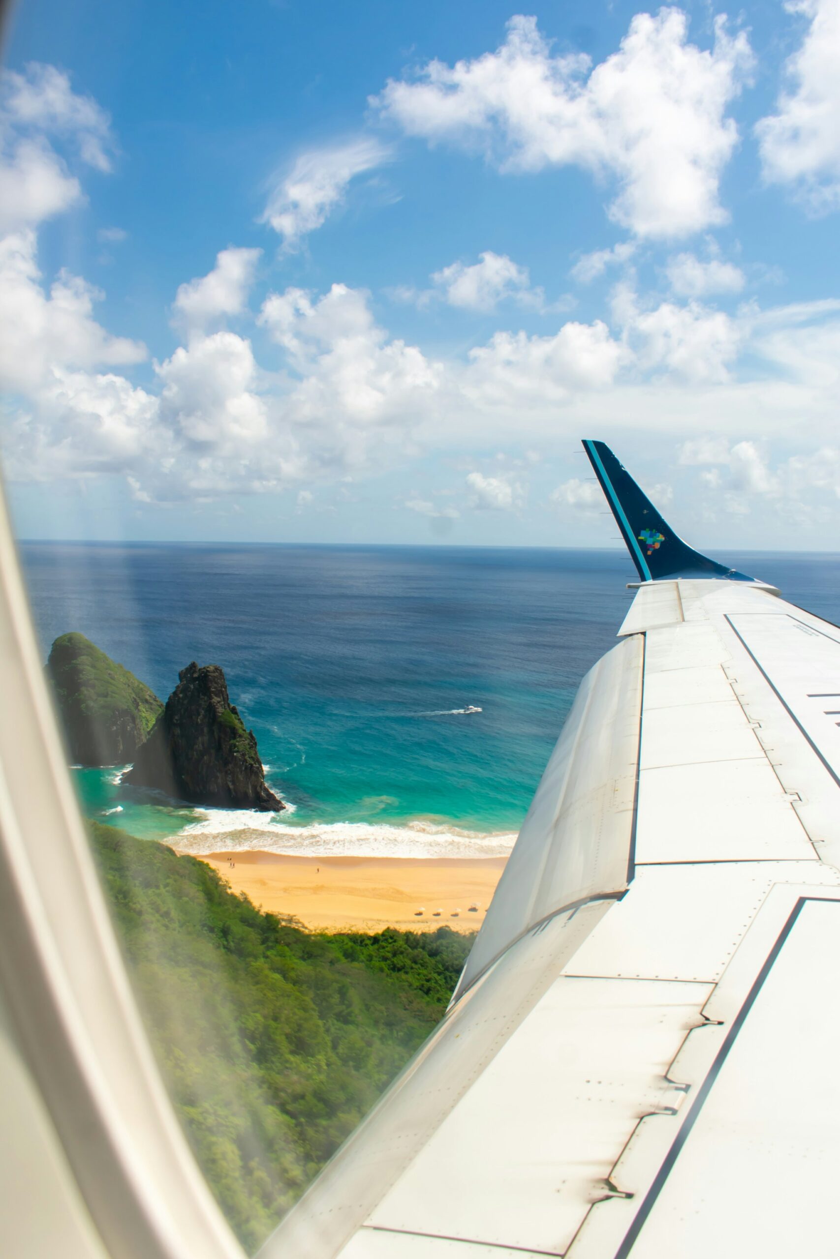 View from an airplane window of a golden beach framed by lush green hills and a dramatic volcanic sea stack, with turquoise and deep-blue Atlantic waters below — the first breathtaking glimpse of Fernando de Noronha on arrival for a 2-Day Diving Package in Fernando de Noronha