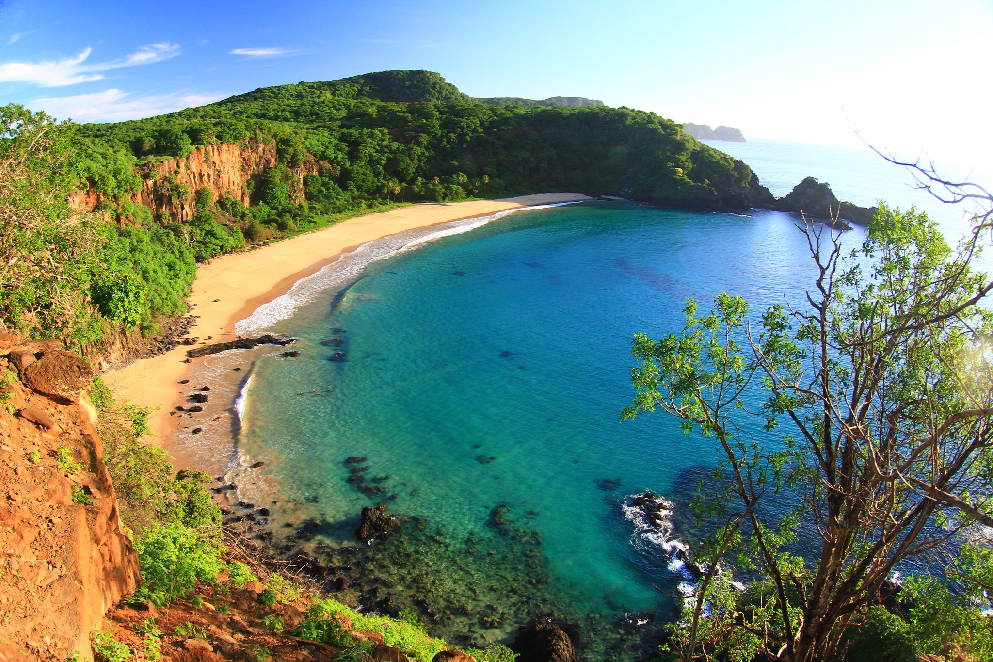 Aerial view of a pristine crescent beach with crystal-clear turquoise water, red sandstone cliffs and dense green forest — one of the iconic bays of Fernando de Noronha, ideal for a 2-Day Diving Package in Fernando de Noronha