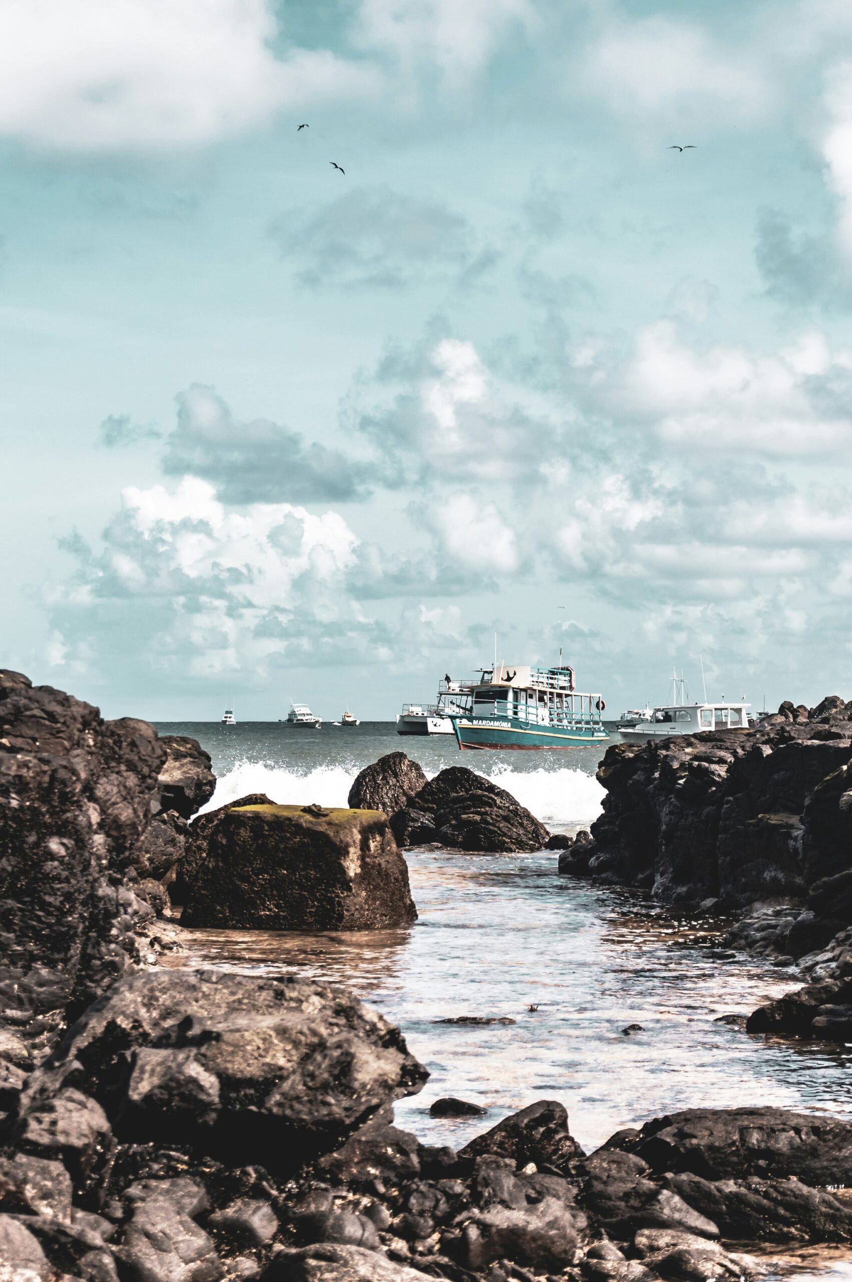 Tour boats anchored beyond a rugged volcanic rock shoreline in Fernando de Noronha, with seabirds soaring above under a dramatic cloudy sky — the departure point for a 2-Day Diving Package in Fernando de Noronha