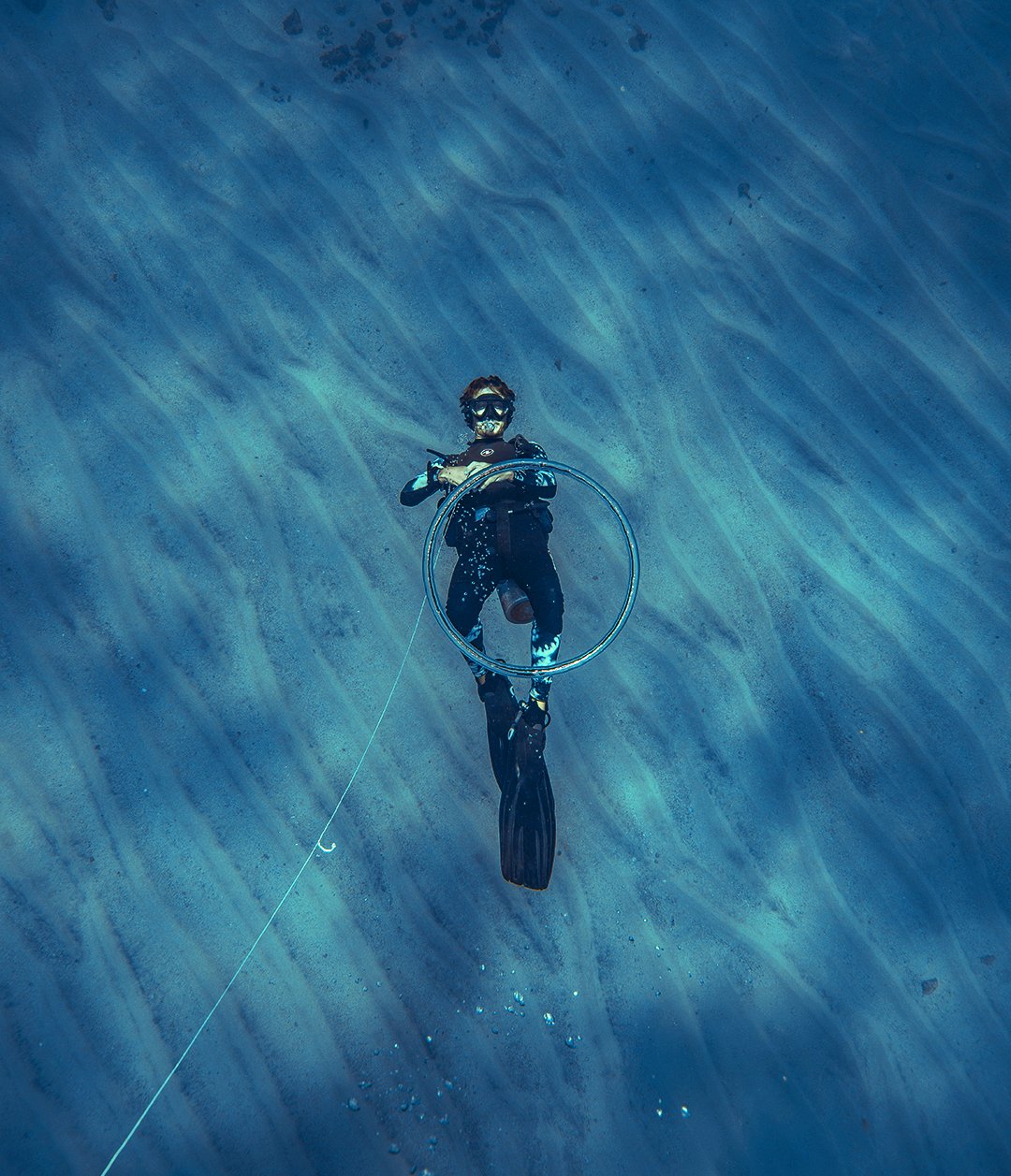 Suspended in blue, a diver drifts above the rippled sand. Aerial view of a scuba diver in a black wetsuit holding a large ring, suspended above a rippled sandy seabed during the 2-Day Diving Package in Fernando de Noronha