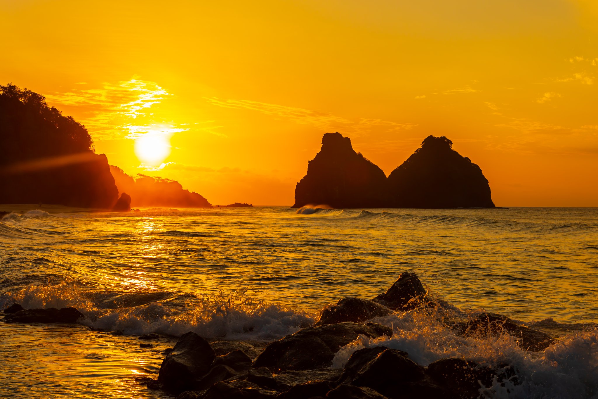 Molten gold poured across the Atlantic — the island's dramatic silhouette at day's end. Photo: Reuber Duarte Fiery golden sunset over silhouetted rocky sea islets off Fernando de Noronha, waves crashing over foreground boulders, part of a 2-Day Diving Package in Fernando de Noronha
