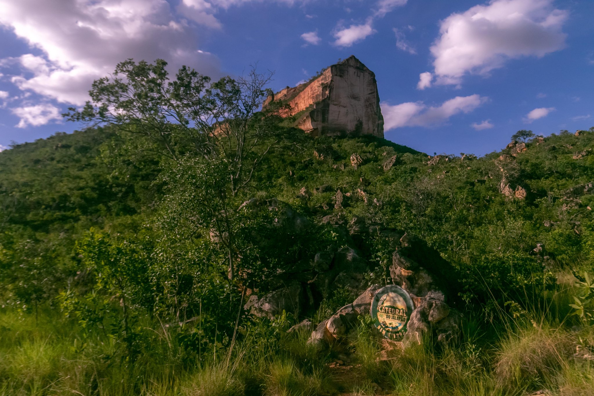 A massive vertical sandstone monolith rises above lush cerrado vegetation on a 4-day Jalapão tour, with a Catedral do Jalapão park sign visible on a boulder in the foreground