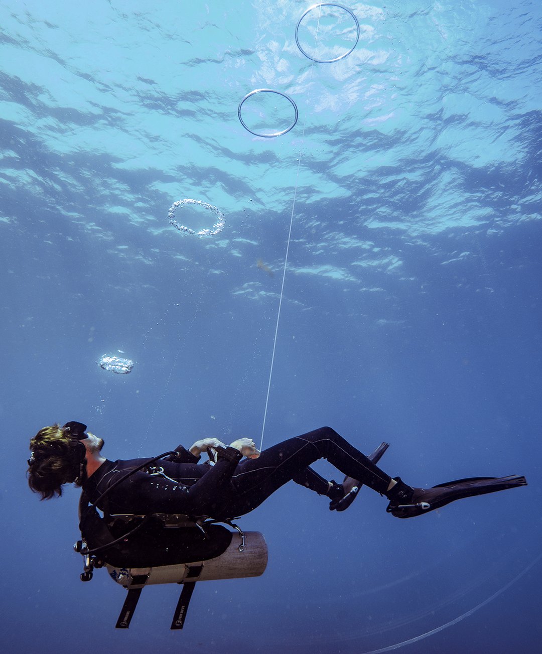A scuba diver in a black wetsuit riding an underwater scooter releases perfect bubble rings that rise toward the sunlit surface during the Adventurer Diving Course in Fernando de Noronha