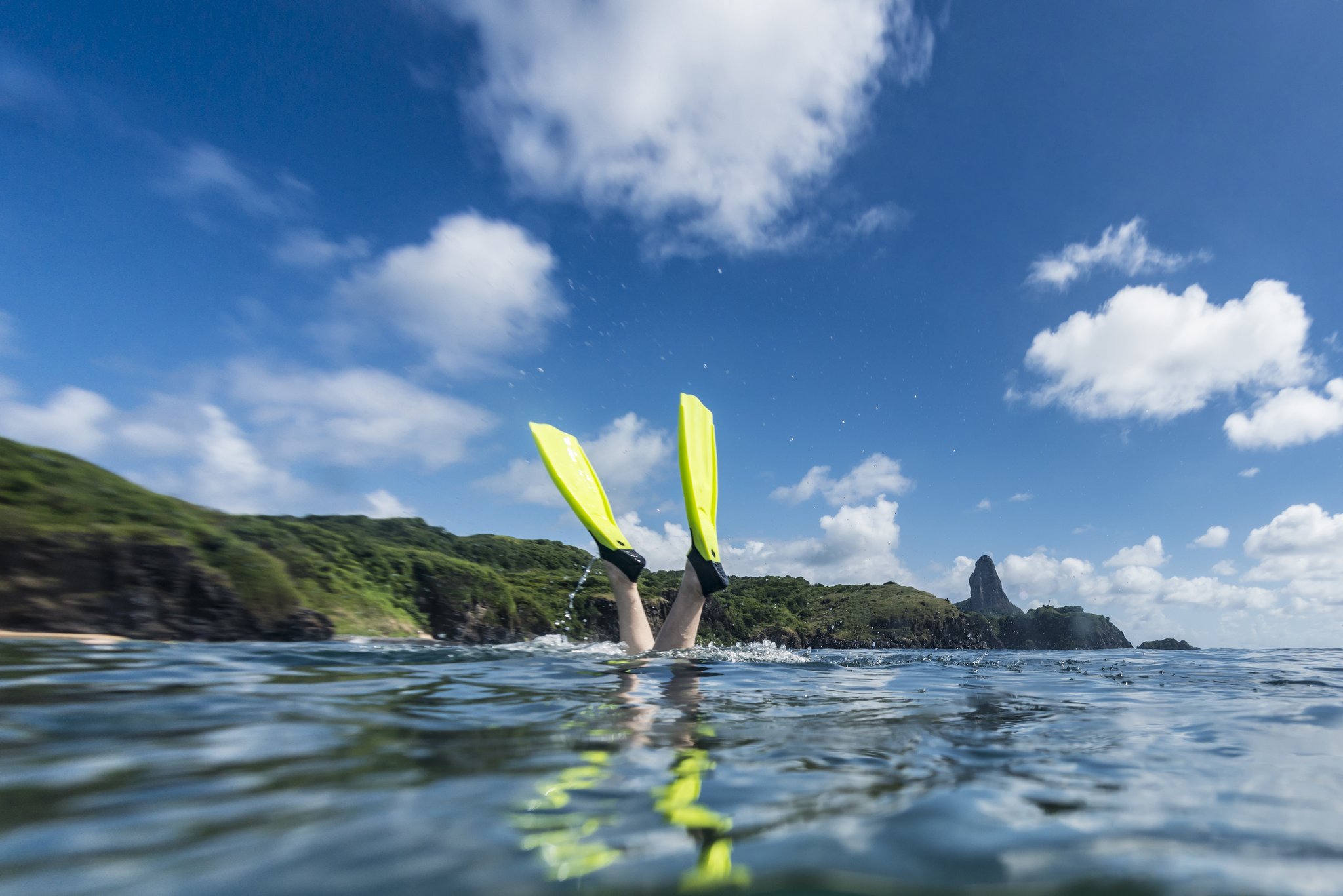 A snorkeler's neon-yellow fins piercing the surface during the Adventurer Diving Course in Fernando de Noronha, with the iconic Morro do Pico peak visible on the horizon