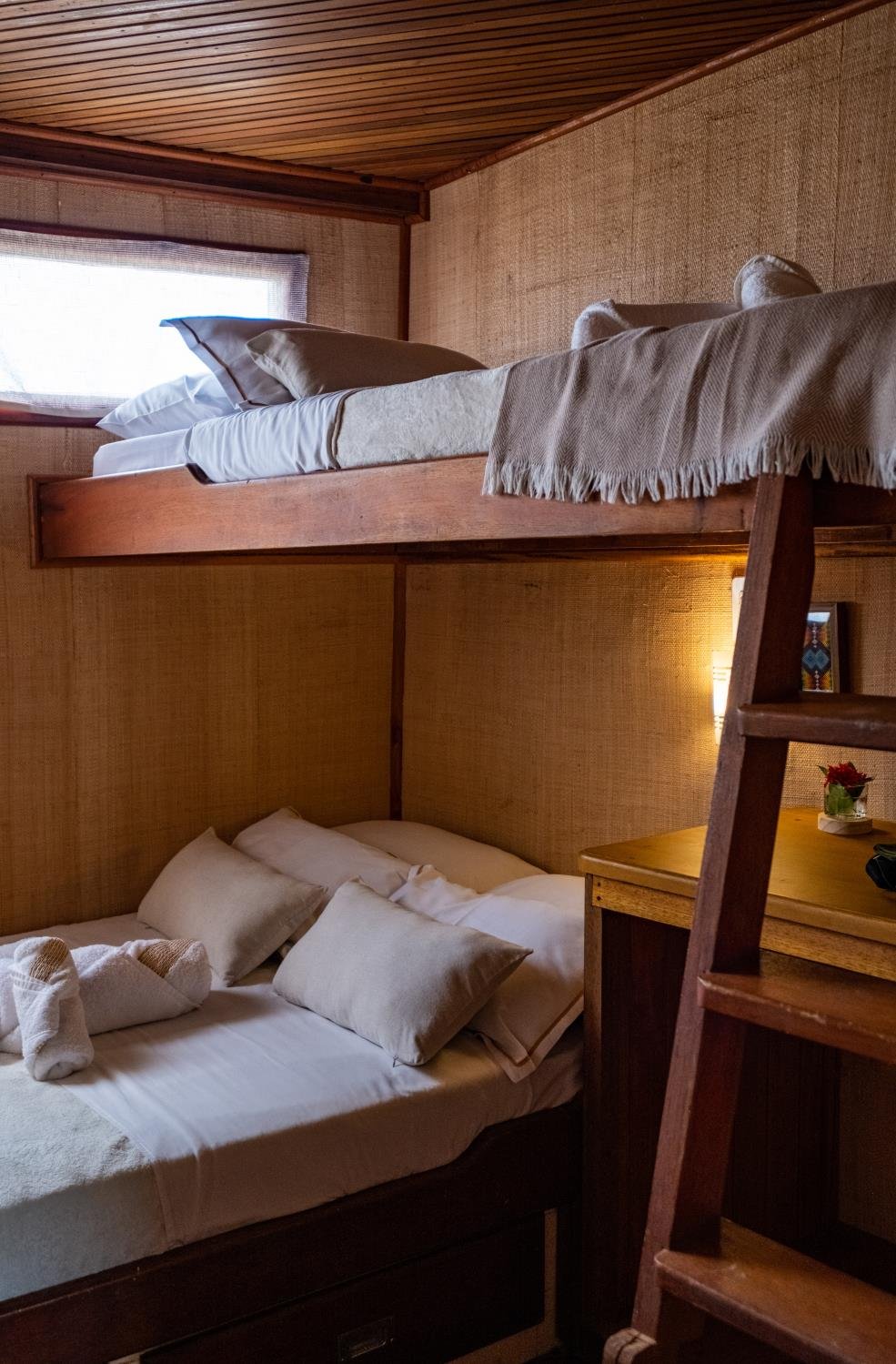 Cosy wooden bunk cabin aboard an Amazon cruise from Alter do Chão, with white linen, cushioned pillows, a fringed throw on the upper bunk, warm bedside lighting, and a timber ladder