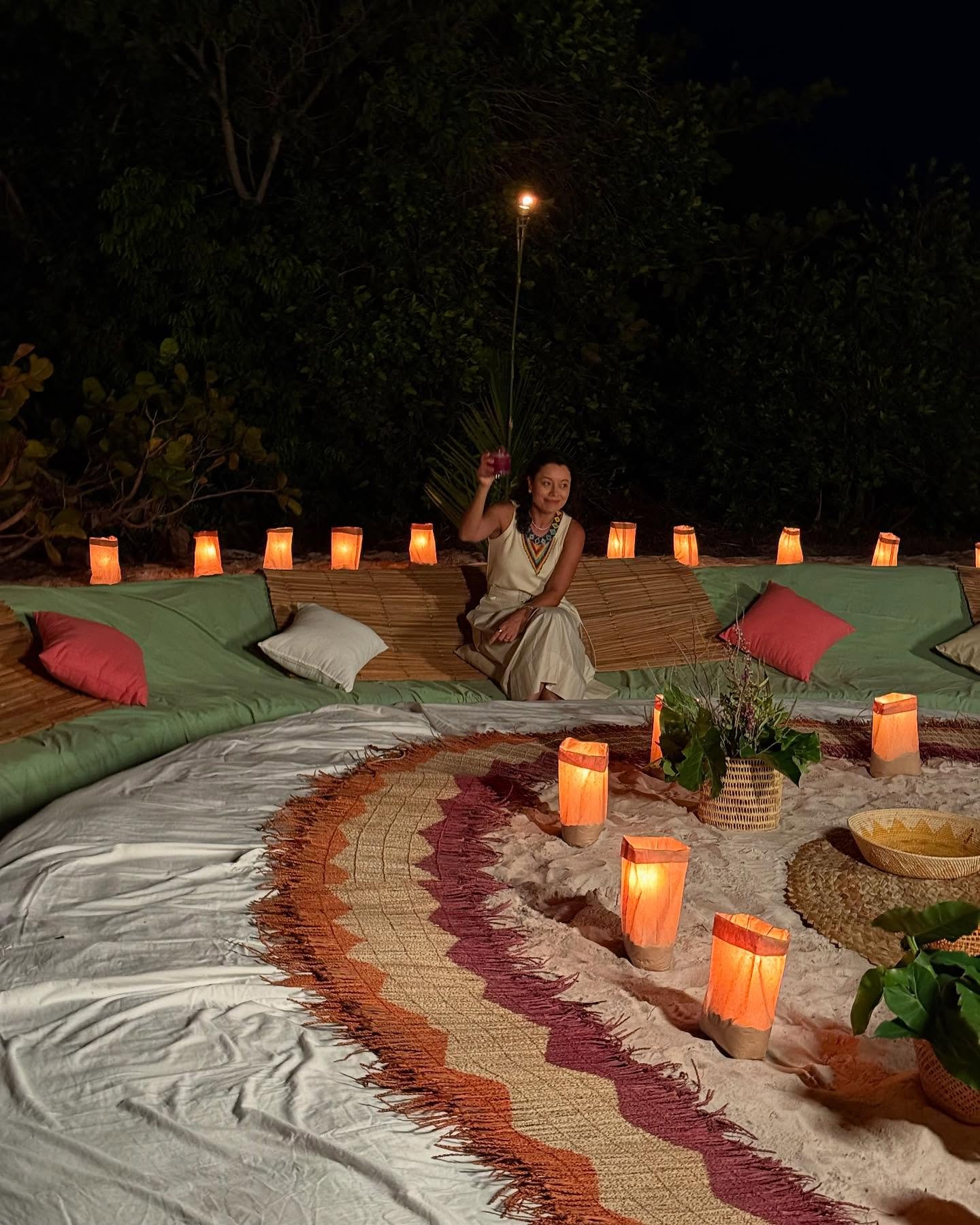 Lanterns in the sand, traditional music on the air — dinner under the Amazon night sky. A woman in a white dress kneels among glowing paper lanterns and woven mats on a sand beach at night, part of an outdoor dinner setup on an Amazon cruise from Alter do Chão