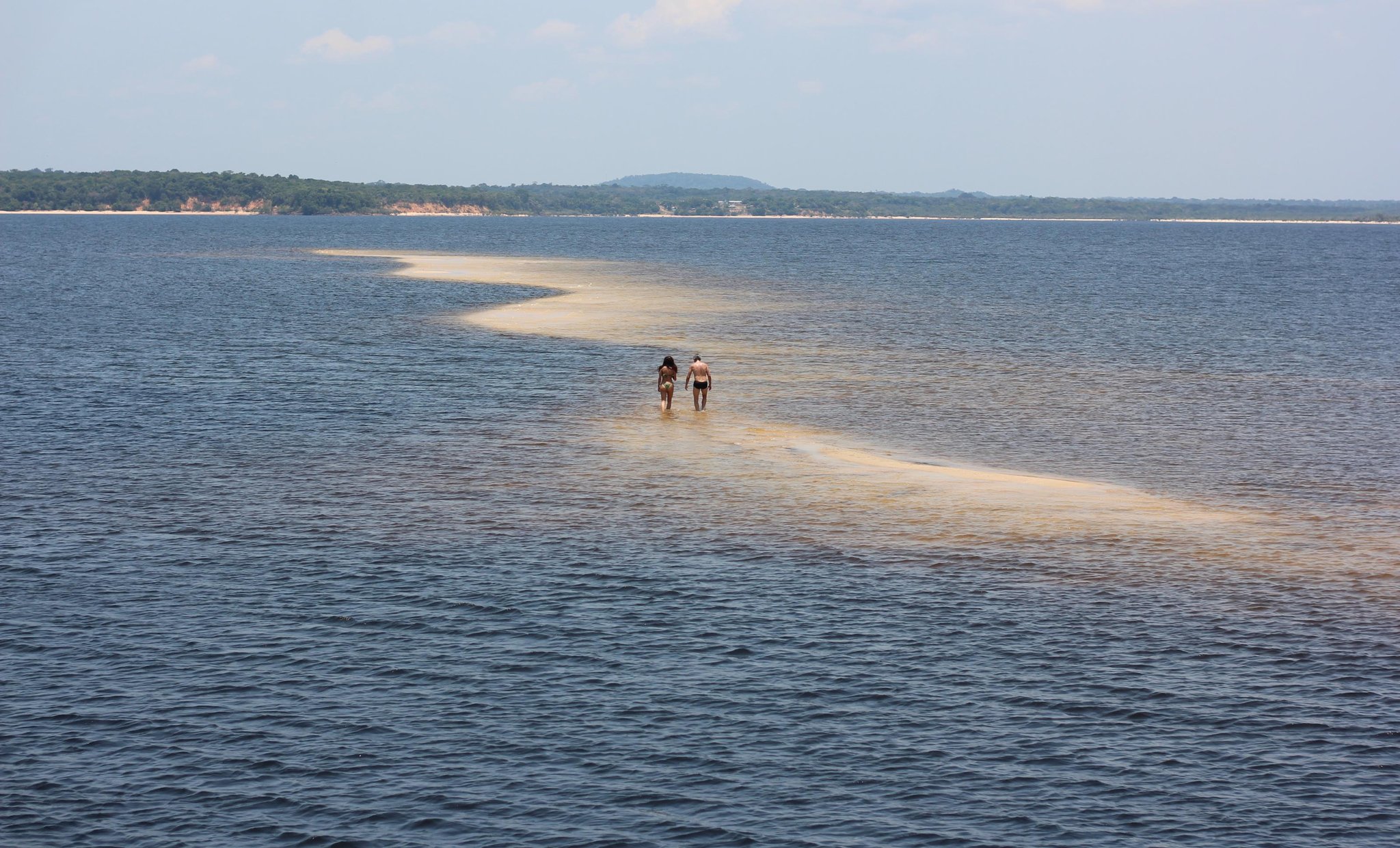 A sliver of sand, a wide river, and nowhere else to be. Two travellers wade along a narrow golden sandbar stretching across wide blue river waters on an Amazon cruise from Alter do Chão, with a forested shoreline and low hills in the distance