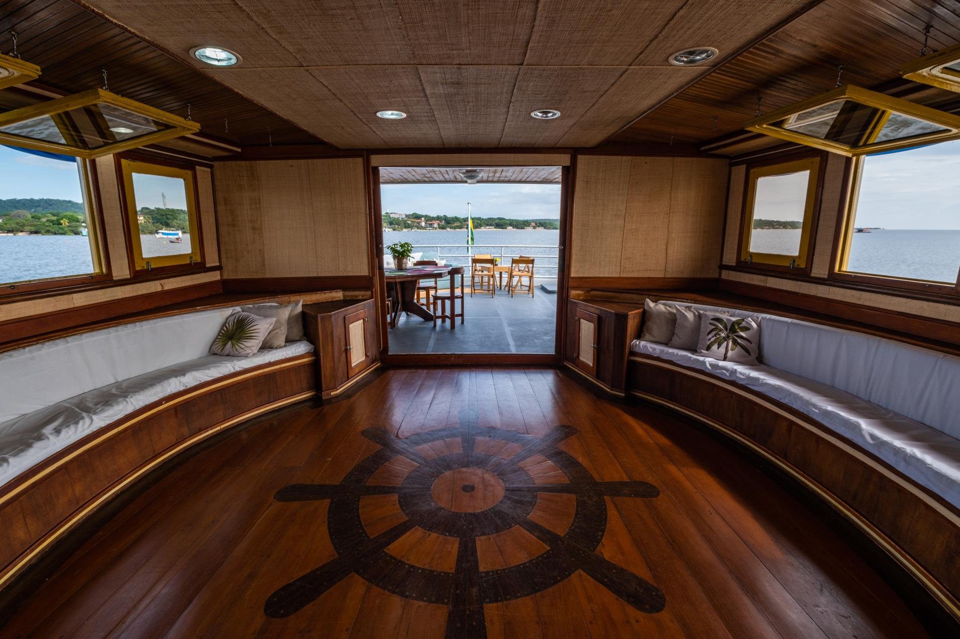 Polished hardwood salon aboard an Amazon cruise from Alter do Chão, with curved bench seating, a ship's-wheel floor inlay and open doors leading to a rear deck overlooking a broad river