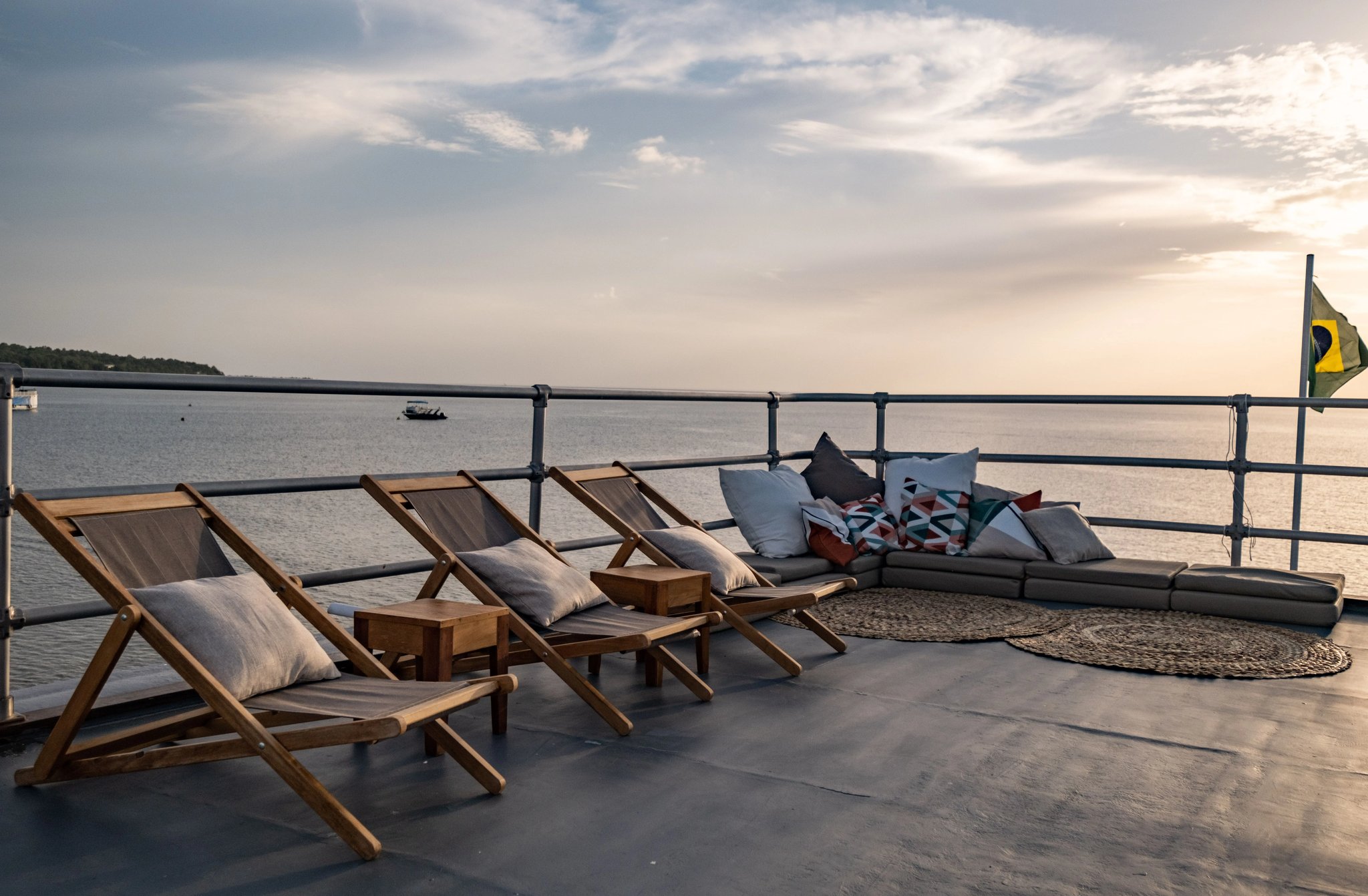 Deck chairs out, river wide, evening almost here. Teak deck chairs and a cushioned lounge corner on the upper deck of an Amazon cruise from Alter do Chão, with a broad river stretching to the horizon under a soft evening sky and the Brazilian flag flying at the stern